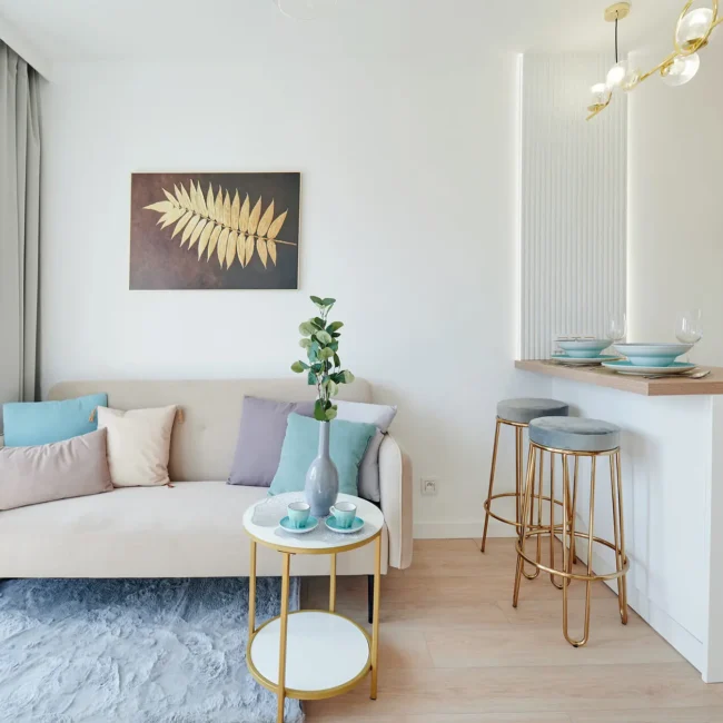 Jak urządzić mieszkanie 25 m2? Nowoczesny i minimalistyczny salon z beżową sofą, złocisto-szklanym stolikiem kawowym, rośliną doniczkową i oprawioną grafiką na ścianie