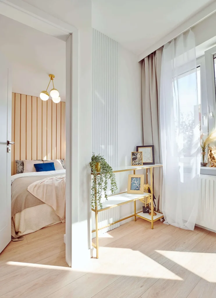 Przytulna i minimalistyczna sypialnia z drewnianą ścianą z akcentami, łóżkiem z beżową kołdrą i małym stolikiem z przedmiotami dekoracyjnymi, takimi jak rośliny i oprawione dzieła sztuki