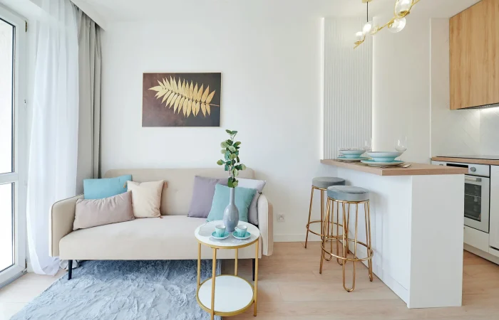 Jak urządzić mieszkanie 25 m2? Nowoczesny i minimalistyczny salon z beżową sofą, złocisto-szklanym stolikiem kawowym, rośliną doniczkową i oprawioną grafiką na ścianie